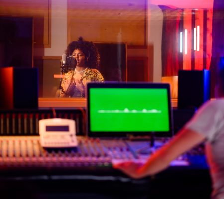 Young talented female singer with curly hair standing in recording isolation room, singing from her heart. Mixing console, monitor, and recording technician in the foreground. Neon light in reflection.